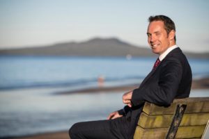 Rangitoto College Principal Patrick Gale sat on a bench, in a relaxed pose and smiling, at the beach, with the sea and Rangitoto Island (Ngā Rangi-i-totongia-a Tama-te-kapua) faded in the background