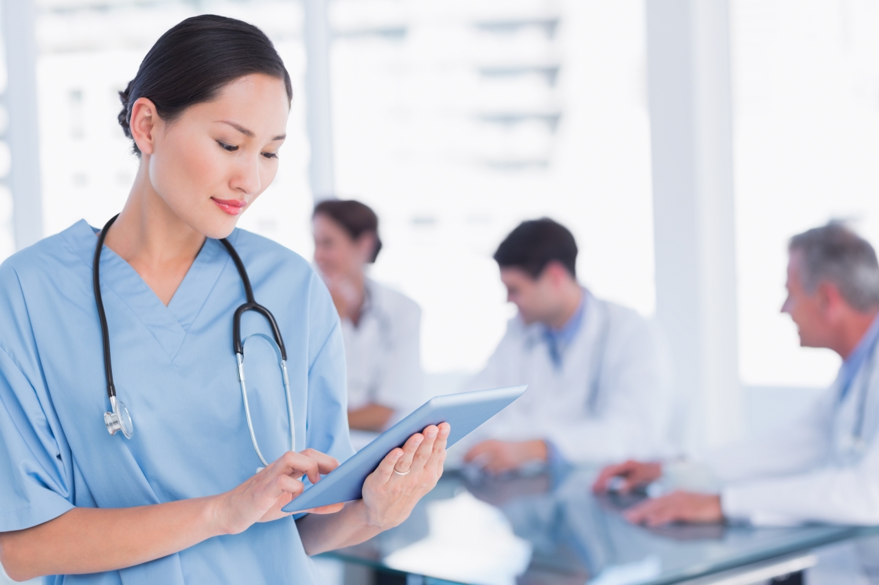 Female medic, wearing a stethoscope around her neck, typing into an iPad with 3 colleagues behind her sat down at a table