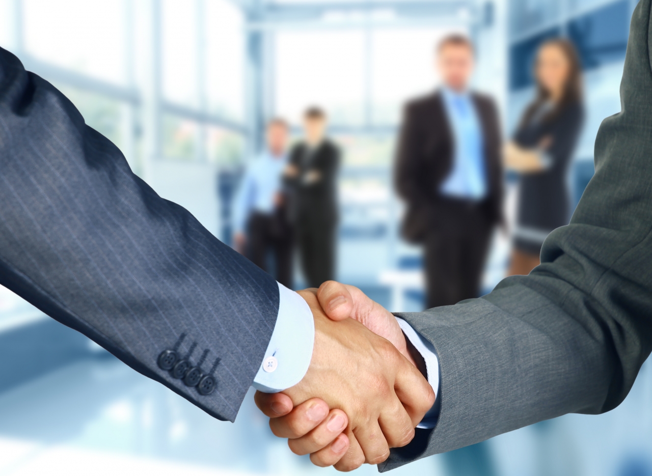 Two men shaking hands wearing suits, in an office, with 3 male colleagues and 1 female colleague faded in background