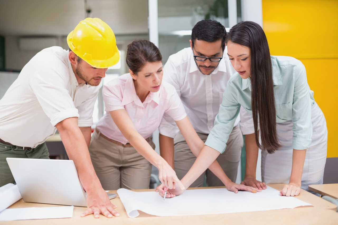 4 engineers, casual dress, 2 male of which one is wearing a yellow hard hat, with 2 females, leaning over a drawing discussing its content