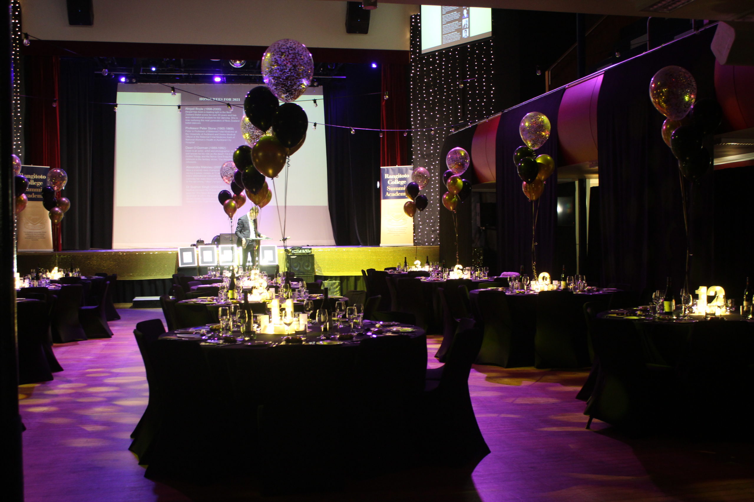 2021 Summit Academy Dinner tables, with lit candles and set of floating gold and black balloons on each table, in Rangitoto College Auditorium