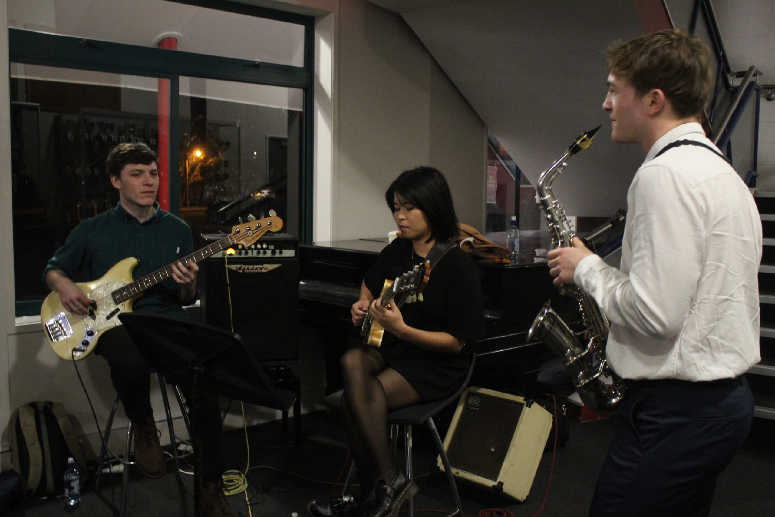 Rangitoto College students playing music, 1 seated male guitarist, 1 seated female guitarist, 1 standing male saxophone player, with a piano in the background, at 2012 Summit Academy Dinner